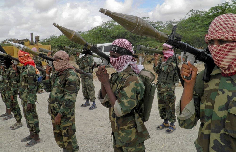 Line of masked soldiers in camouflage uniforms aiming shoulder-fired weapons with large rocket launchers in a dusty outdoor area.