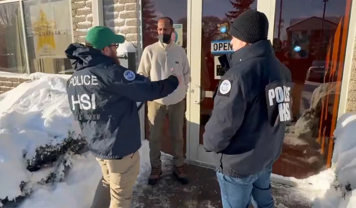Two officers in HSI jackets speak with a man at a store entrance on a snowy sidewalk, near a glass door with an 'OPEN' sign visible.